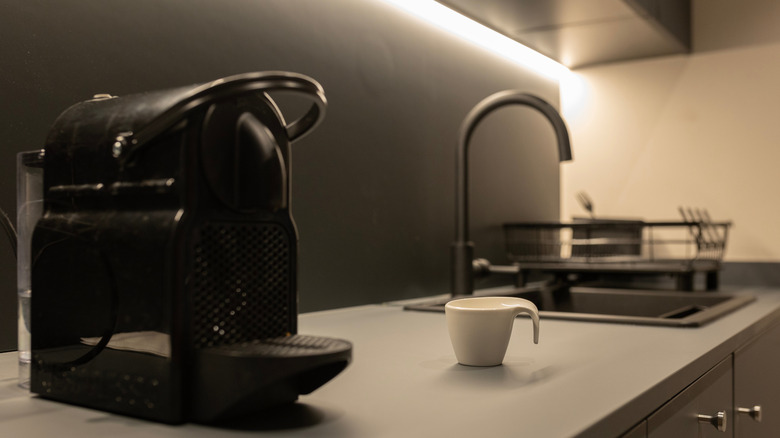 A kitchen counter with a black coffee maker on it. A with coffee mug and a sink are in the background.