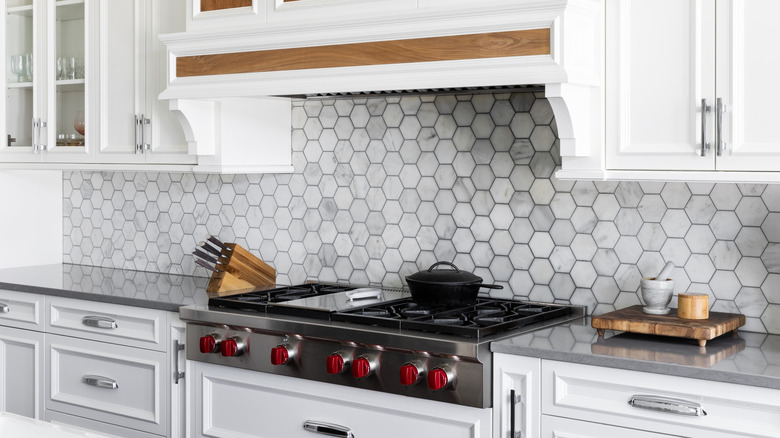 A kitchen range detail with a marble hexagon tile backsplash, white cabinets, stainless steel stovetop and range hood, and gray countertop.