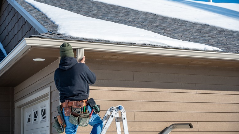 Person on a ladder inspecting gutters with some snow on the roof