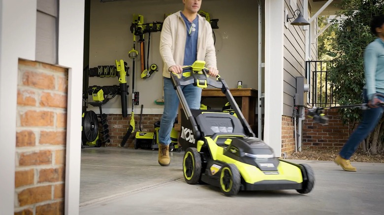 Man pushing Ryobi lawn mower out of garage with other Ryobi yard tools