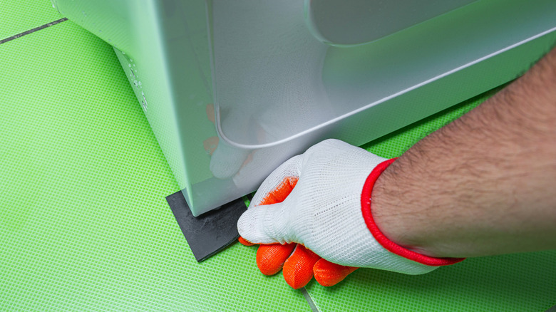 A gloved man's hand placing rubber under a washing machine to balance it.