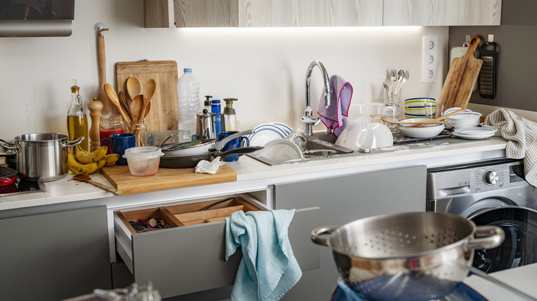 A messy kitchen with dirty dishes, dish cloths, and utensils piled on counters
