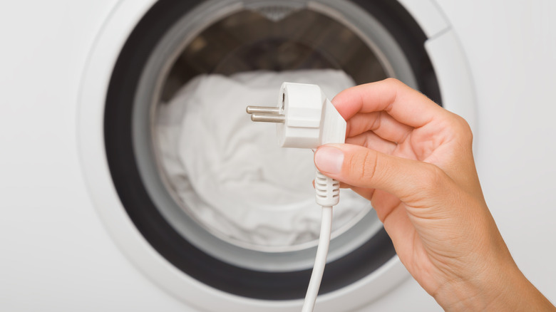 A hand holding an electrical cord in front of a front-loading washing machine.