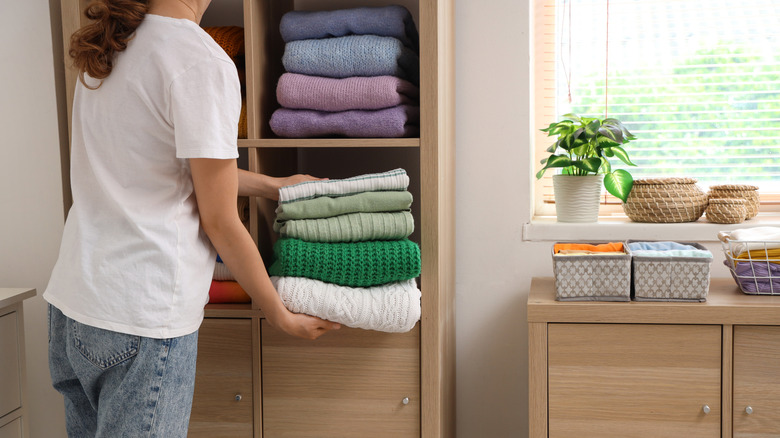 A woman neatly stacking green jumpers into a closet space. A pile of purple and blue jumpers is in the compartment above.