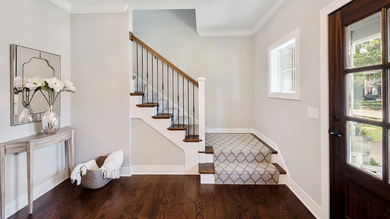 An entrance porch with hardwood floors and cream walls. There is a small table with a vase and large white flowers in front of a mirror and a basket with blankets on the ground.