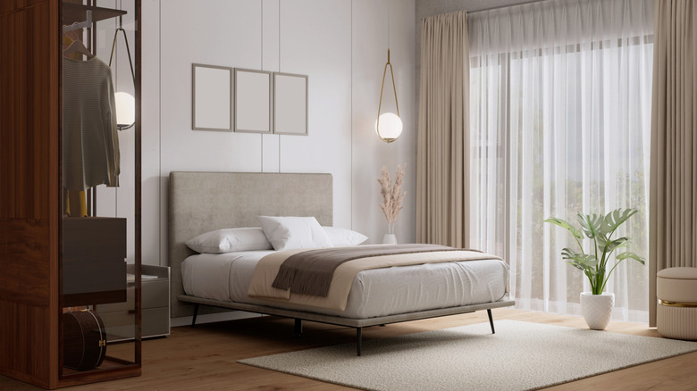 A modern bedroom with beige bed, cream curtains and a large green plant in a pot on the floor.