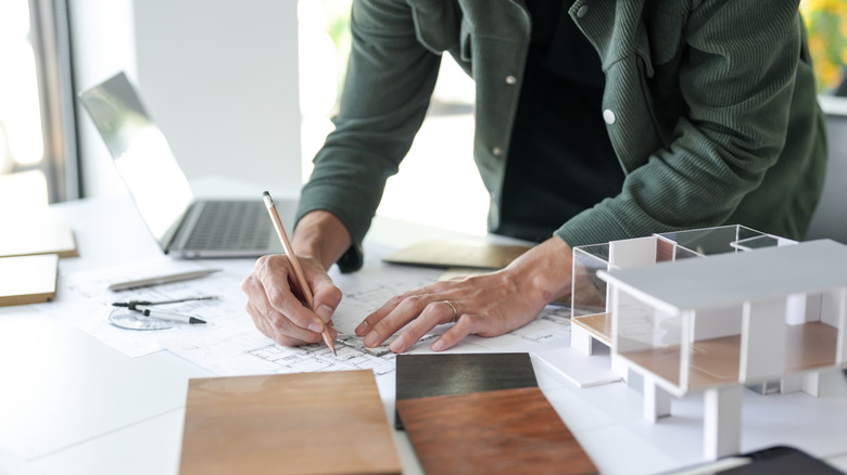An architect at a desk full of materials and models draws a kitchen floor plan on paper.