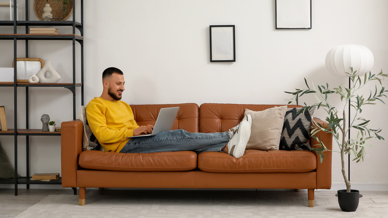 A man using his laptop sits on a brown leather couch in the living room.