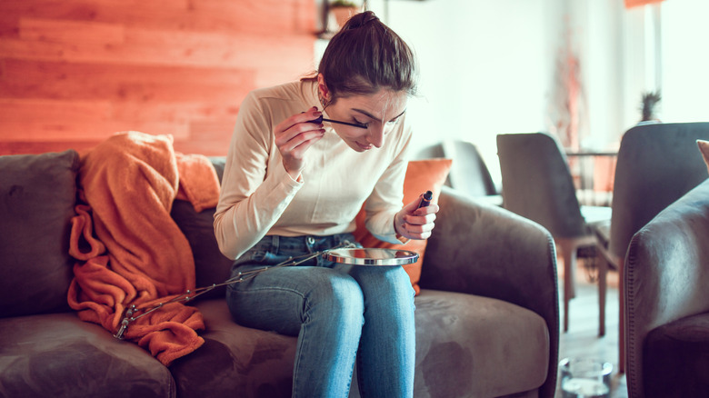 A woman applies mascara while sitting on a suede couch.