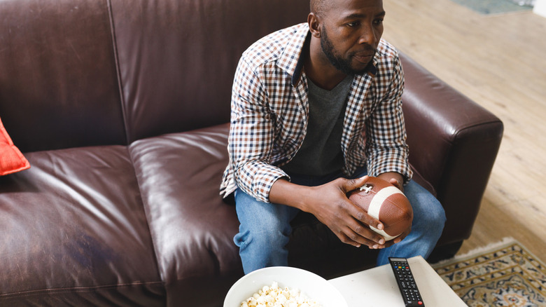 A man holding a football sits on leather couch while wearing blue jeans.