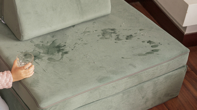 Water spilled onto a green suede couch from a glass of water held in a hand.