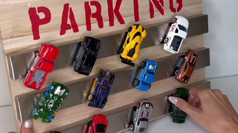 Woman's hand is placing metal toy cars on a magnetic knife holder on a board that says PARKING in red letters