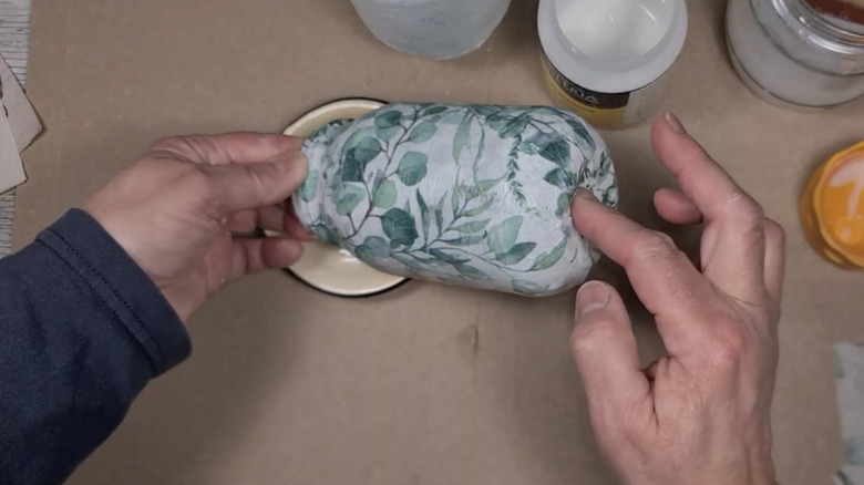 Two hands hold a decoupaged glass jar over a tabletop covered in crafting supplies.