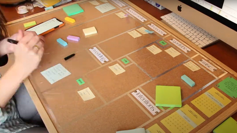 A person writing on a corkboard repurposed as an organizer with sticky notes and tape stuck unto it