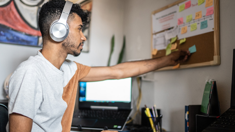 A young man is shown wearing headphones while attaching a small sticky note to a corkboard on the wall near his desk.