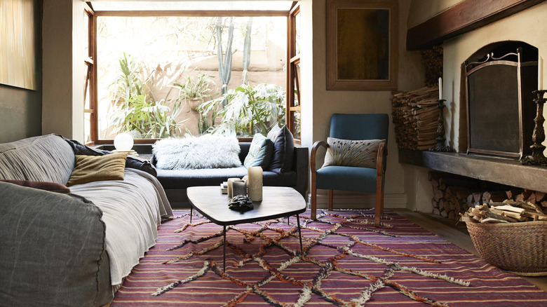 A living room with a big open window, a fireplace, a grey couch, a coffee table, and a woven mauve rug.