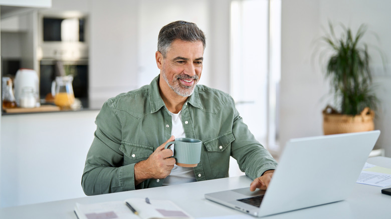 Older man scrolling on laptop while drinking from coffee mug