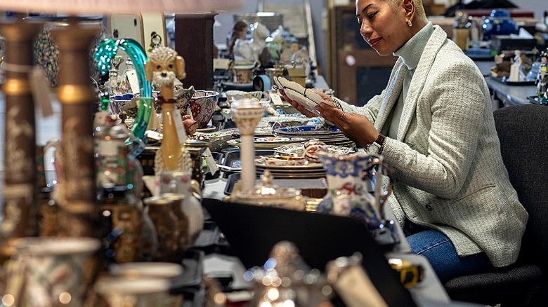 A person inspects a plate while sitting down at a table of antiques