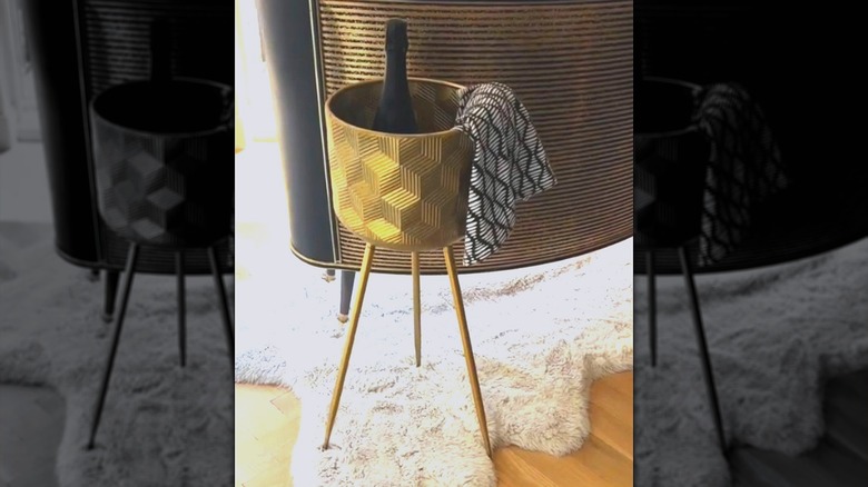 A gold planter stand holds a bottle of wine and a dish towel instead of a plant