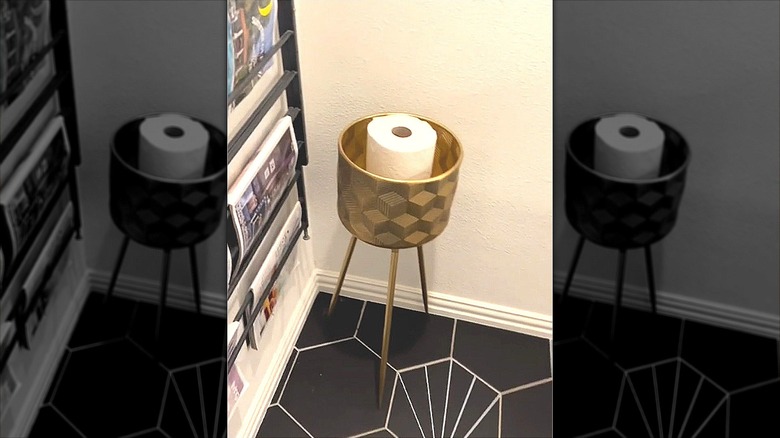 A gold pot planter stand holds toilet paper rolls in a bathroom.