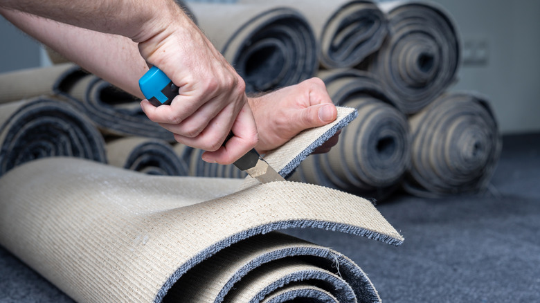 Hands cut blue carpet to fit a space with rolls of carpet in the background