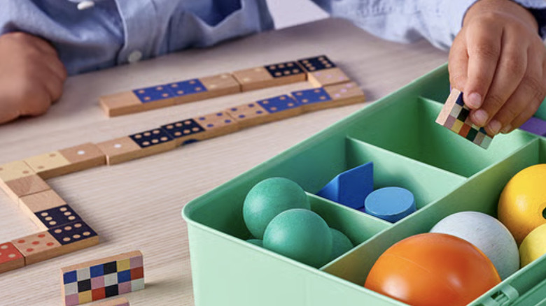 A child puts a domino into a large green IKEA GLIS box with dividers filled with rubber balls and building blocks.