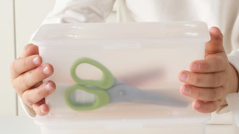A person holds a clear IKEA GLIS container with a pair of green-handled safety scissors inside.