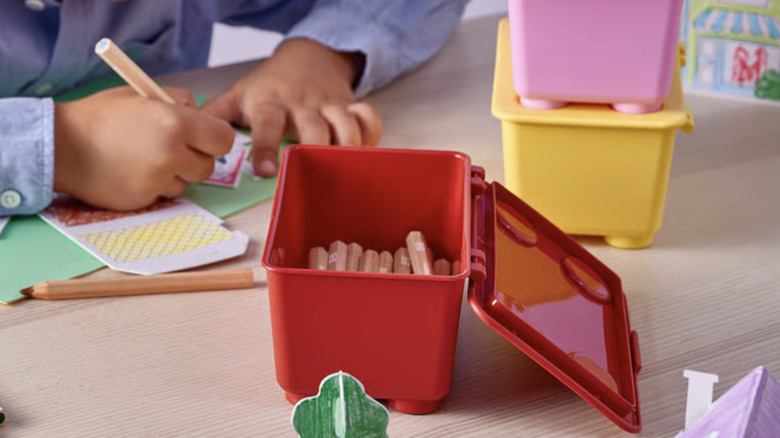 A red IKEA GLIS box holds colored pencils with other boxes and a child drawing on paper in the background.