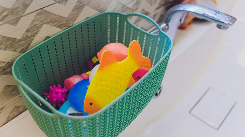 Bath toys in a green plastic basket beside the tub in a bathroom.