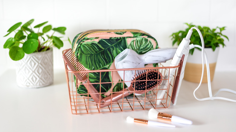 A small pink wire basket with a hair brush, comb, blow dryer, and flat iron.