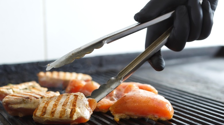 person in latex gloves handling chicken pieces on grill with metal tongs