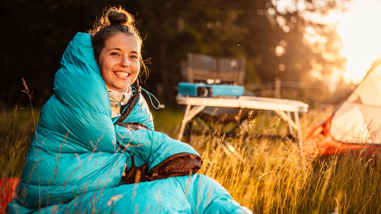 A smiling woman wrapped up in a sleeping bag outside