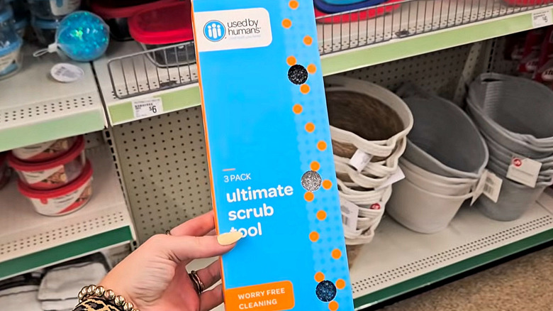 A person with manicured nails holds up a blue package of scrubbers with Dollar Tree shelves in the background