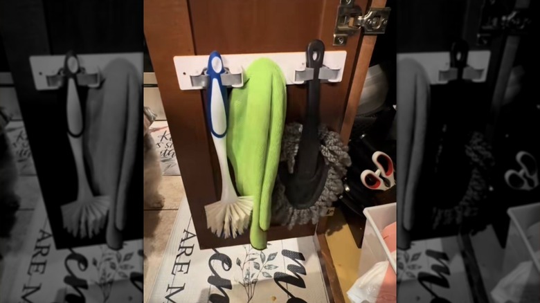 A Dollar Tree Broom Holder on the back of a cabinet door holding cleaning supplies.