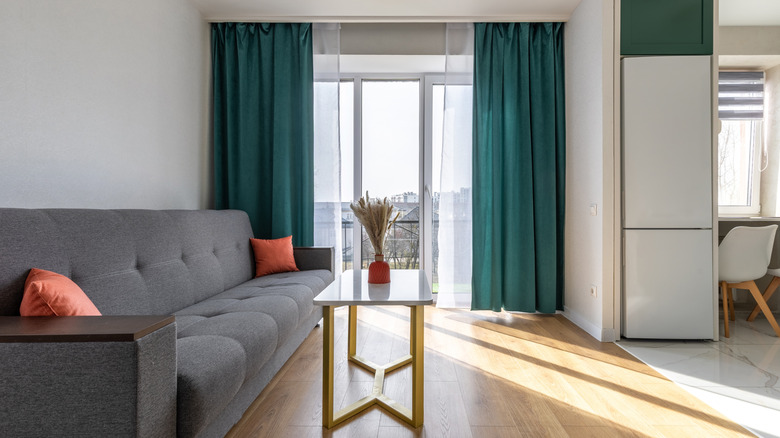 A modern living room with a large gray sofa, a stylish marble coffee table, a red vase with dried flowers, and floor-to-ceiling windows with green curtains.