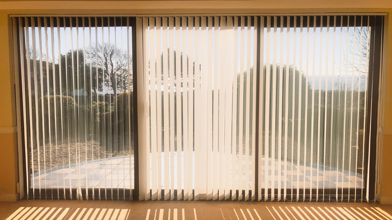 sunlight shining through full length white vertical blinds in front of three glass sliding french doors leading to a patio, garden and sea view, creating shadow stripes.