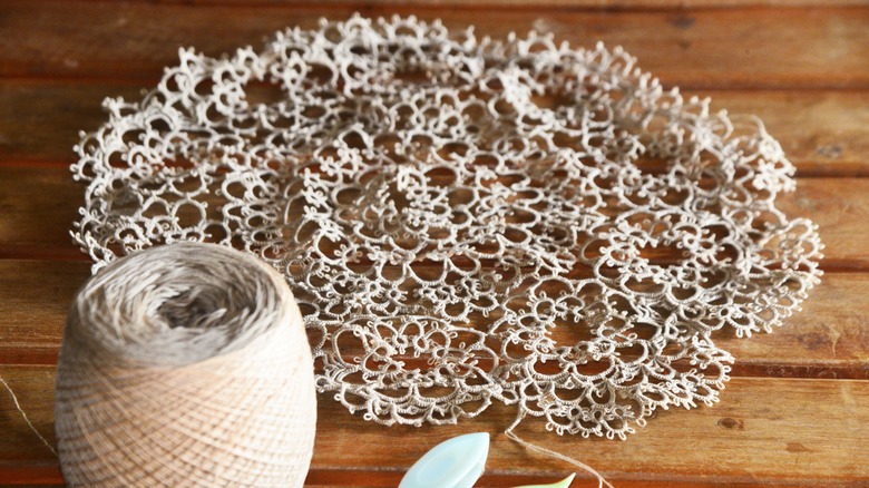 A round piece of tatted lace sitting next to a spool on a wooden, slatted surface.