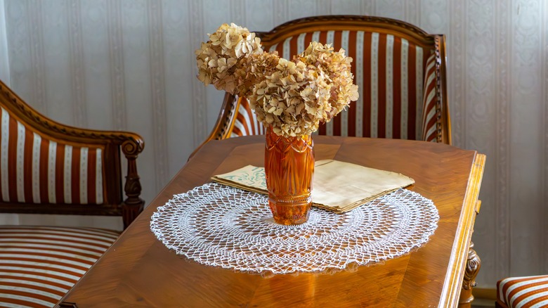 A vintage doily on an antique table set with a vase of dried hydrangeas sitting on top of it.
