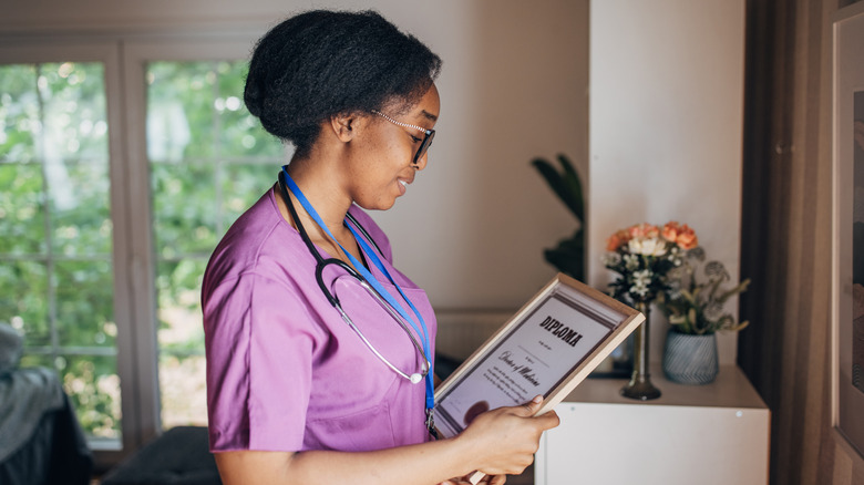 A woman holds framed certificate while wearing purple scrubs and stethoscope.
