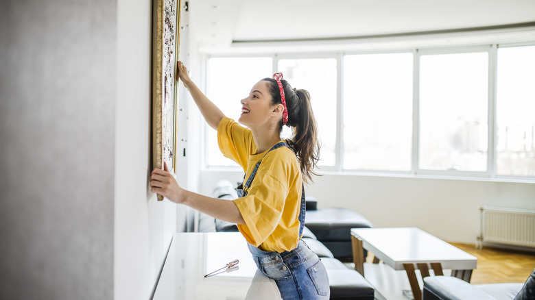 A woman in a yellow t-shirt hangs art on wall.