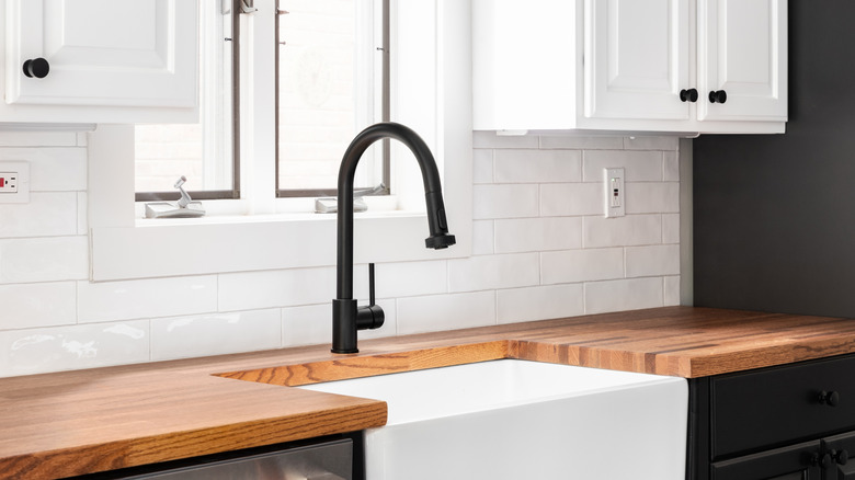 Dark butcher block counter with white farmhouse sink, black faucet, and white backsplash.