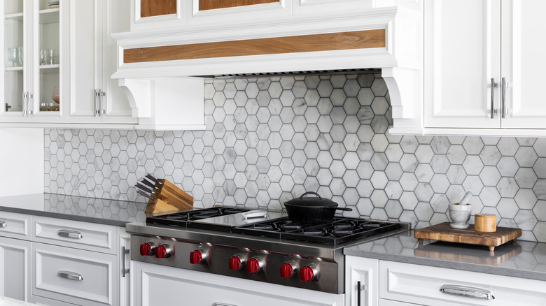 A kitchen range detail with a marble hexagon tile backsplash, white cabinets, stainless steel stovetop and range hood, and grey countertop.