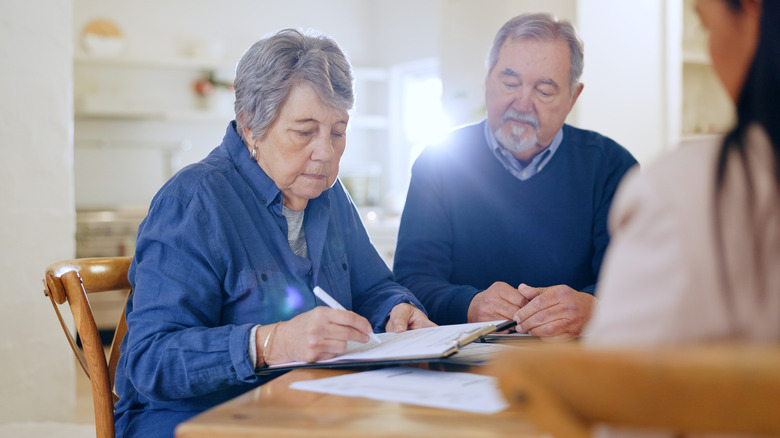 An older woman is singing some documents while her husband looks on