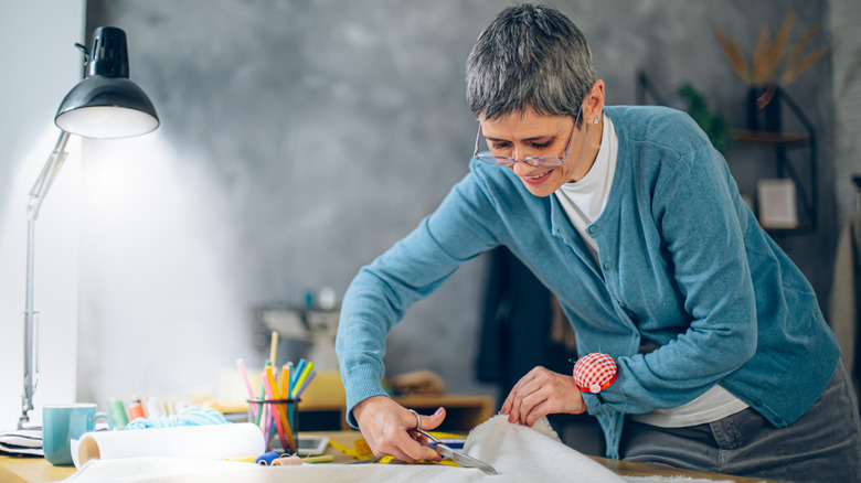 person cutting fabric with a desk lamp, tea mug, and thread on the table