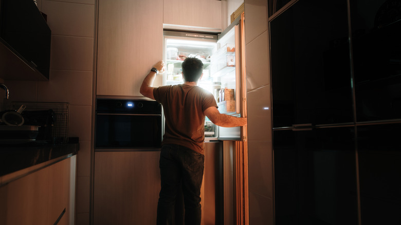 A man stands in front of an open refrigerator in a dark kitchen
