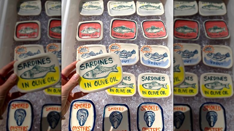A hand holding a sardine-themed plate in front of a display of other fisher-themed plates