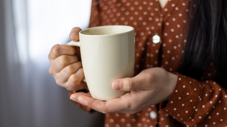 A woman in a brown button down shirt holding an off-white coffee mug.