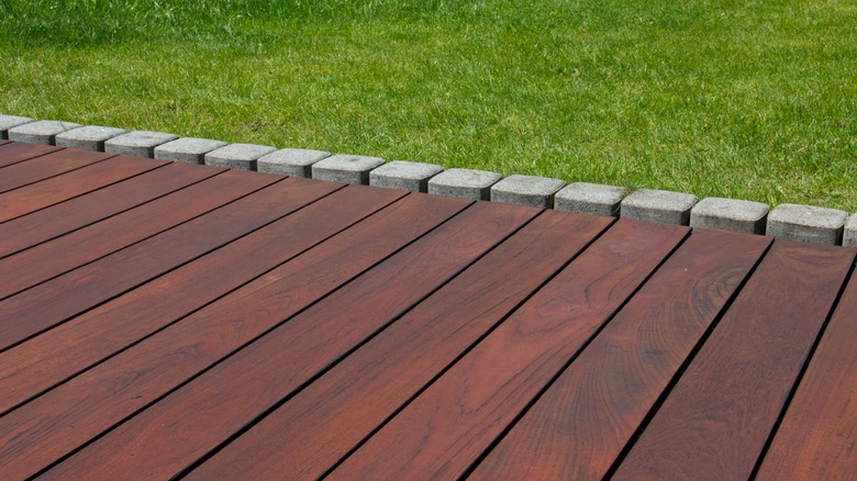 A dark colored wooden deck bordered by square stones and green grass.