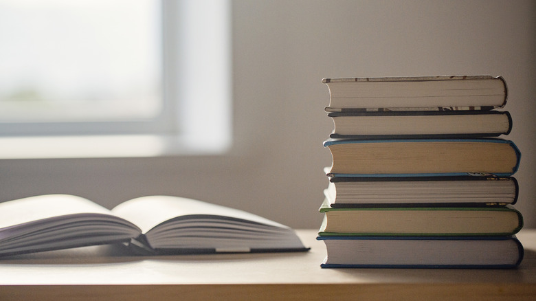A stack of assorted books sitting next to and open book on a wooden desk with sunlight coming through the window.