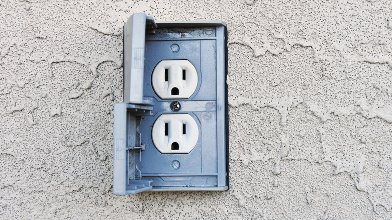 An outdoor outlet with two plugs and safety cover on a textured grey wall